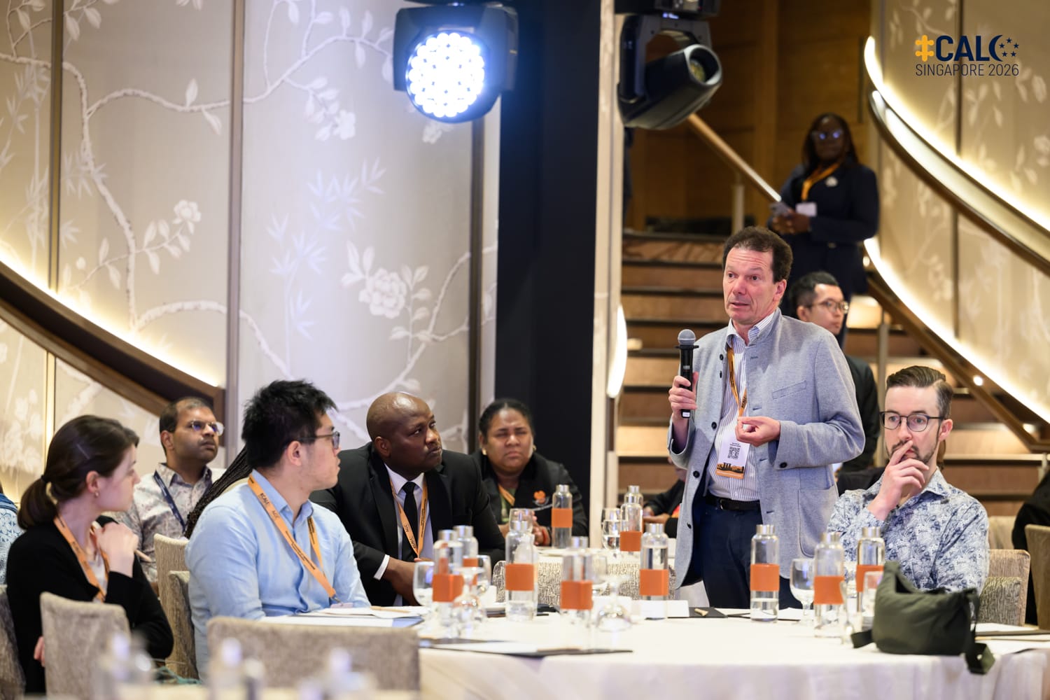 A delegate stands to ask a question during a Conference session at The Fullerton Hotel Singapore, with other delegates seated at the round table listening on.